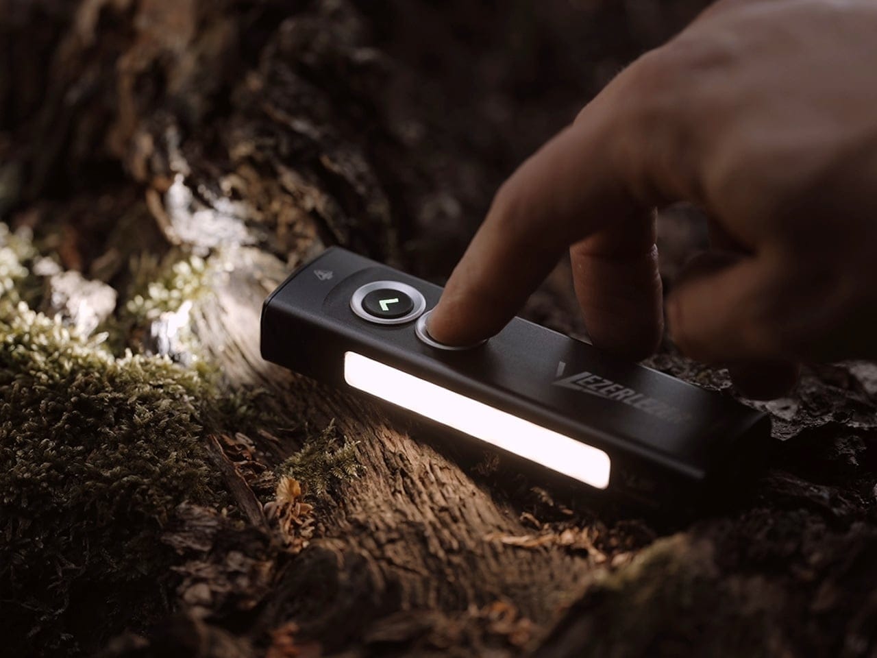 Close-up of a finger pressing a button on a small rectangular outdoor LED light resting on a mossy log in a forest.